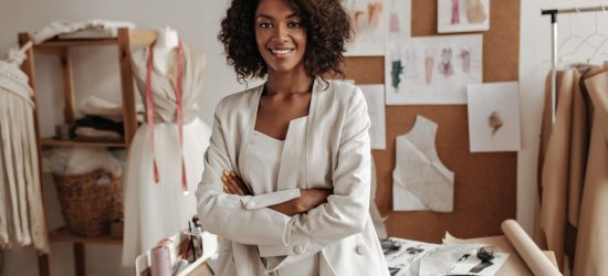 Beautiful curly brunette dark-skinned fashion designer poses in office, leans on table. Young lady in white suit crosses arms and smiles.