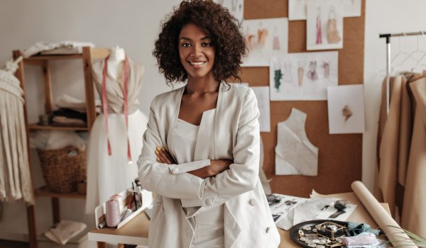 Beautiful curly brunette dark-skinned fashion designer poses in office, leans on table. Young lady in white suit crosses arms and smiles.