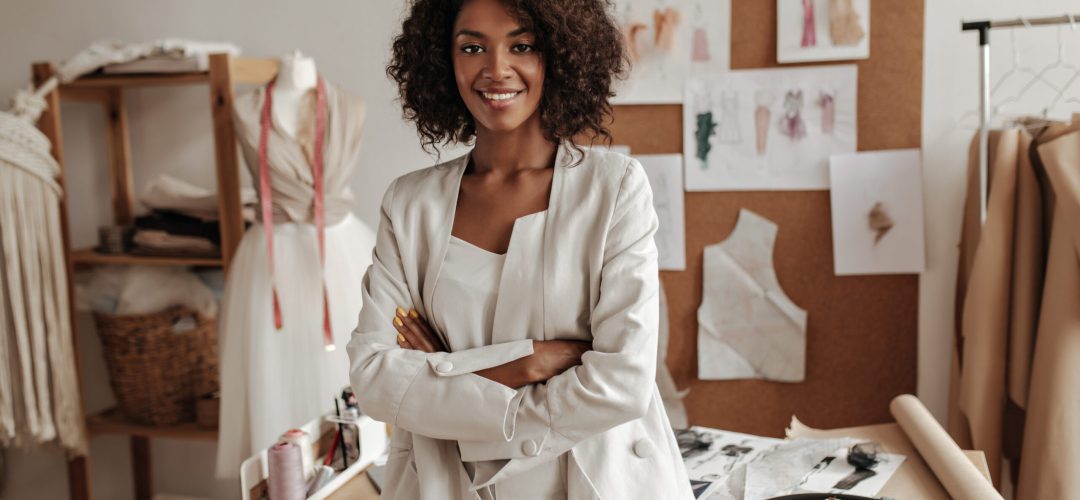 Beautiful curly brunette dark-skinned fashion designer poses in office, leans on table. Young lady in white suit crosses arms and smiles.